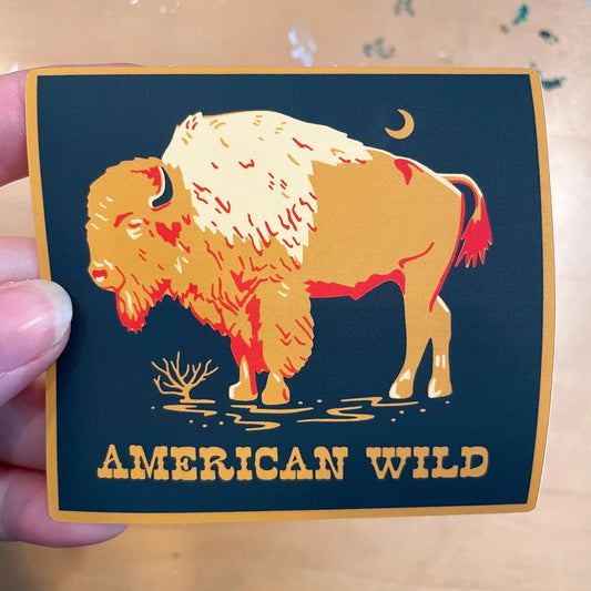 Square shaped 3.5” by 3” vinyl sticker. Mustard yellow border. Navy background. Mustard yellow bison with red and cream details is in the center of the sticker. The bison is standing in a puddle. There is a small crescent moon in the right corner above the bison. Western style text “American Wild” is below the bison. 